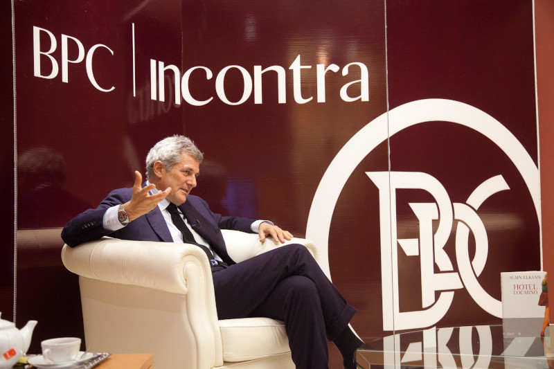 Alain Elkann parla seduto su un divano bianco, con un logo BPC sullo sfondo.
