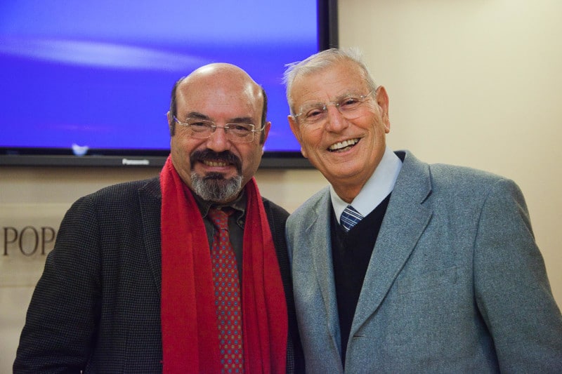Due uomini sorridenti in un evento, uno con sciarpa rossa e l'altro in giacca grigia.