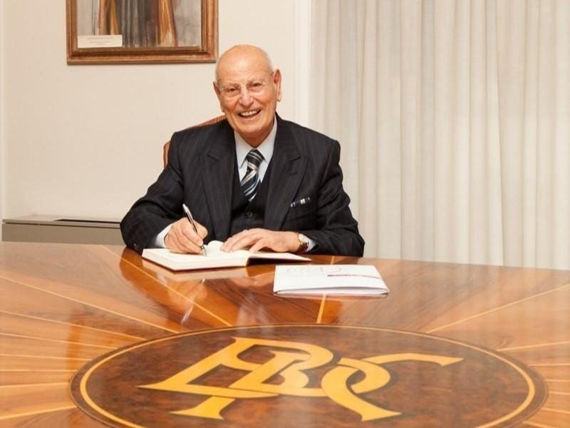 Un uomo anziano sorridente, seduto a un tavolo con un libro aperto, mentre scrive.