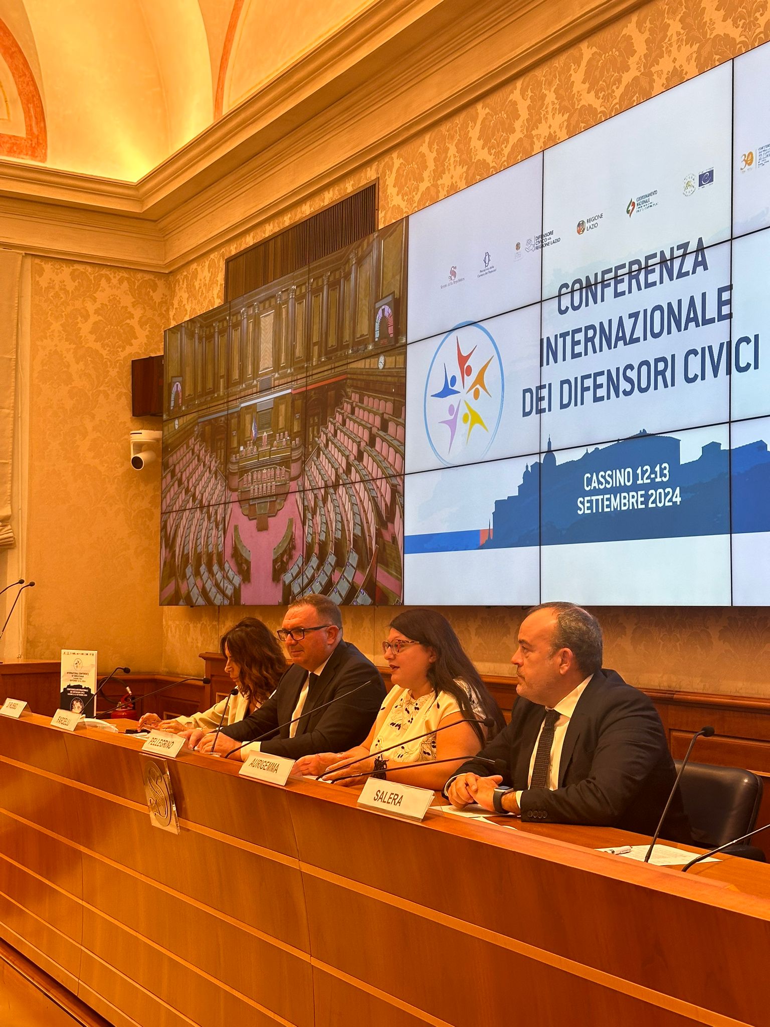 Conferenza Internazionale dei Difensori Civici a Cassino, con relatori al tavolo e schermo con logo dell'evento.