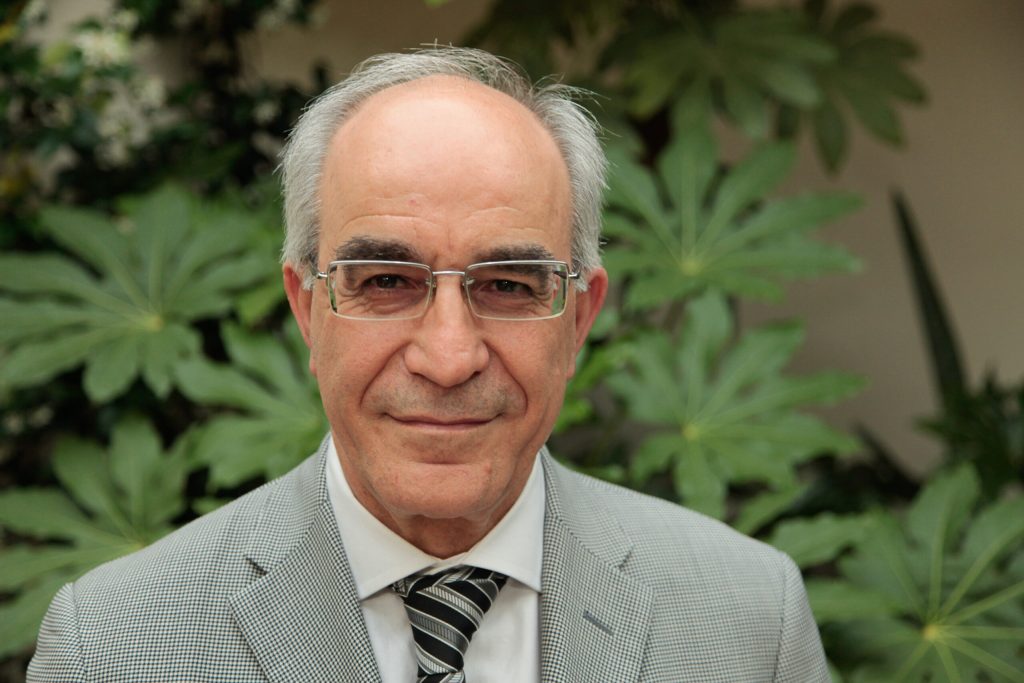 Giovanni Pacitto, ex Vicedirettore Generale, in giacca grigia con camicia e cravatta, sorridente davanti a piante verdi.