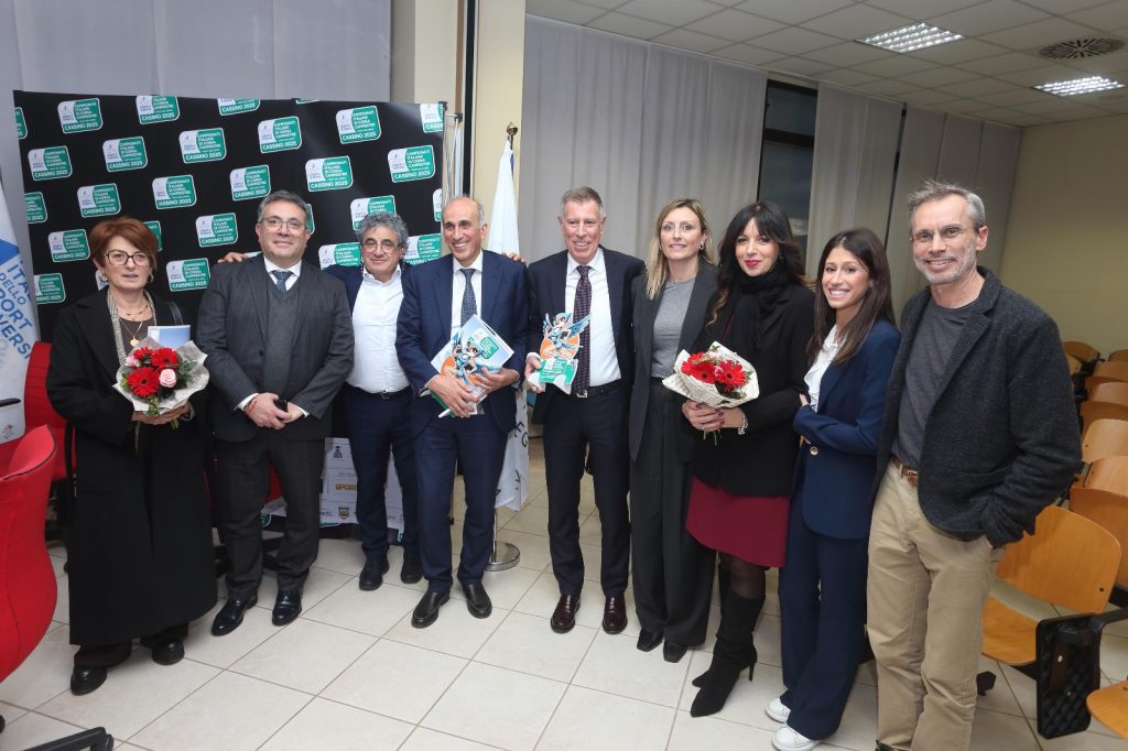 Gruppo di persone sorridenti in un evento, con fiori in mano e un premio, in un ambiente interno.