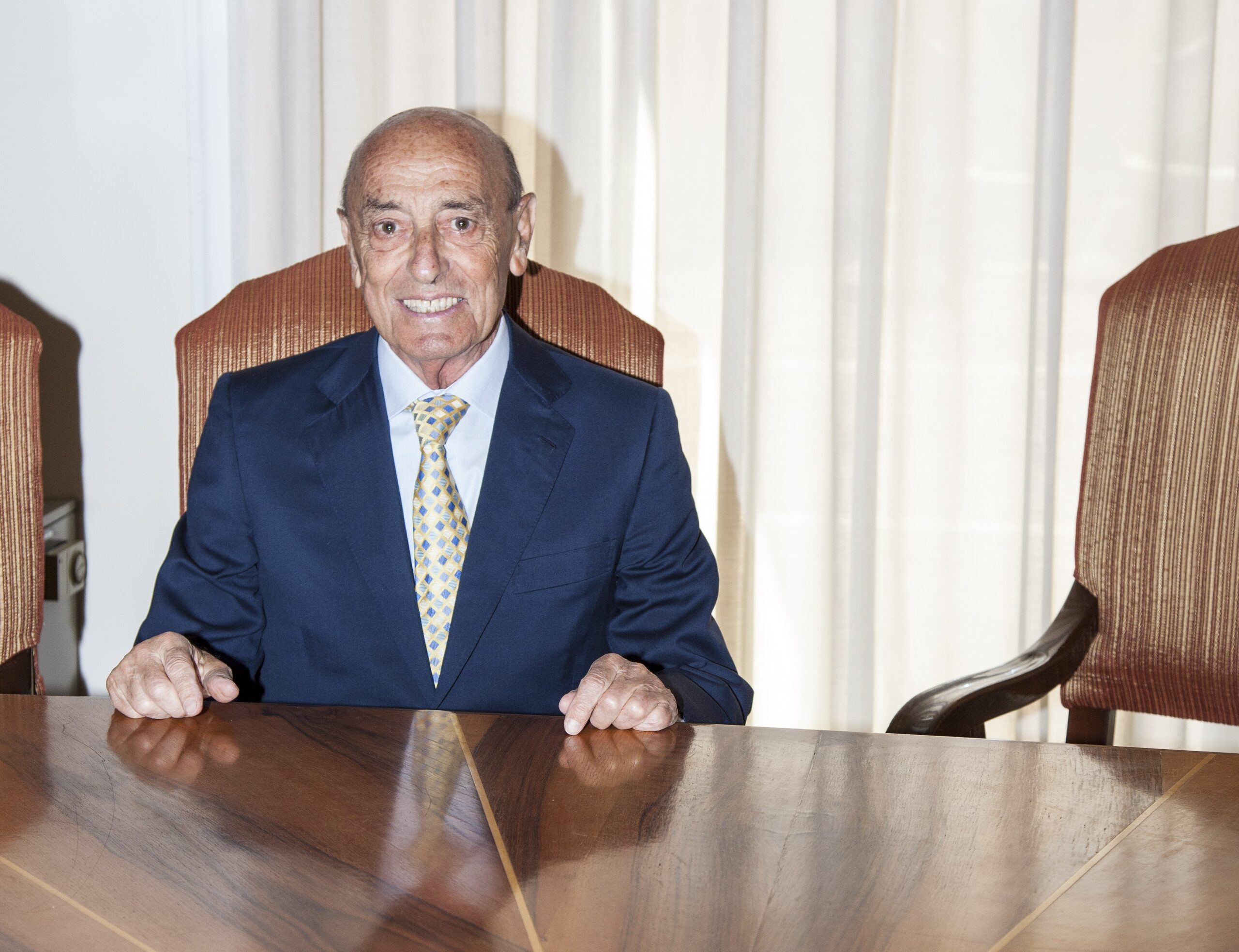 Emilio Giangrande sorridente seduto a un tavolo, indossa un abito blu e una cravatta a fantasia.